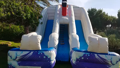 מגלשה מתנפחת גדולה עם שני מסלולים ודפנות כחולות בעיצוב של התזות מים, הממוקמת בחוץ בין עצים ודשא.