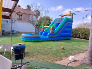 מגלשת מים מתנפחת גדולה בצבע כחול עם עצי דקל, המוצבת על הדשא בחצר אחורית שטופת שמש.