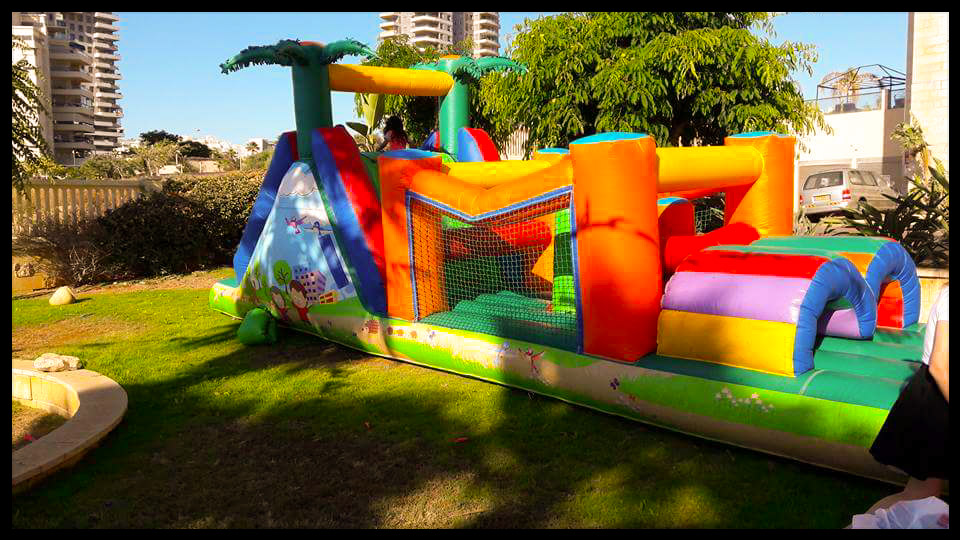 מתקן קפיצות מתנפח וצבעוני עם מגלשות ומנהרות, המוצב על מדשאה ירוקה תחת שמיים שטופי שמש.