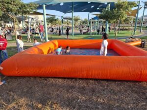 ילדים משחקים בבריכה מתנפחת קטנה בצבע כתום בפארק, כשברקע נראים מתקני משחקים.