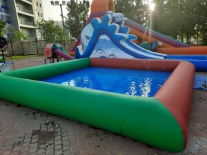 בריכה מתנפחת קטנה עם מים צלולים, לצד מגלשה מתנפחת צבעונית על משטח לבנים בחוץ.
