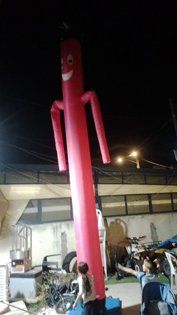 בלילה, איש צינור מתנפח גבוה ואדום עם פרצוף מחייך עומד בחוץ ליד אנשים וציוד.