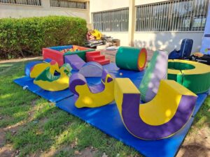 צורות משחק רכות וצעצועים בצבעים עזים מסודרים על מחצלות כחולות בחוץ, ליד בניין ושיחים.