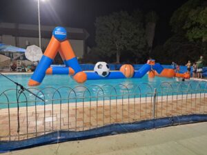 מסלול מכשולים מתנפח עם כדורגל צף בבריכה בשעות הלילה, ואנשים צופים מהגדה.