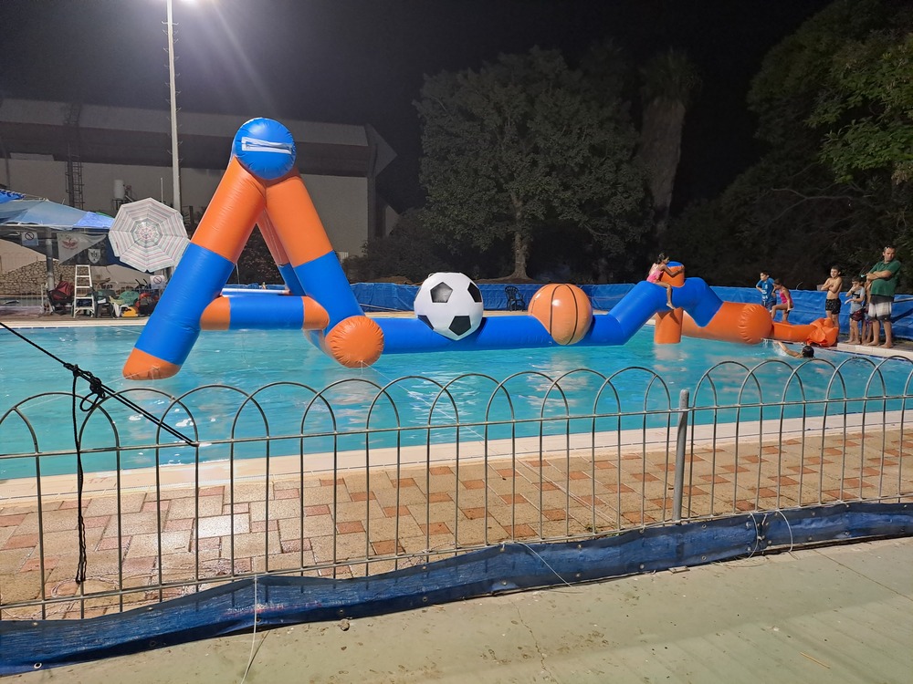 מסלול מכשולים מתנפח עם כדורגל צף בבריכה בשעות הלילה, ואנשים צופים מהגדה.