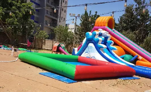 מגלשת מים מתנפחת וצבעונית שהוקמה בחוץ על משטח מרוצף, כשברקע נראים עצים ומבנים.