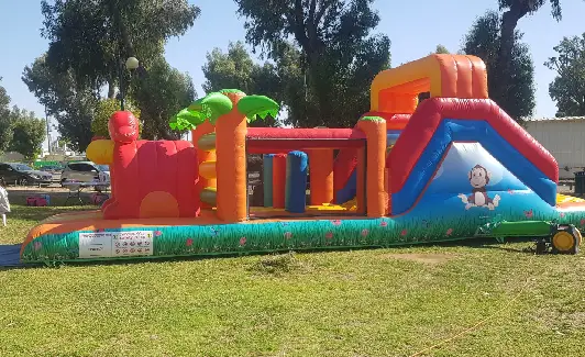 מסלול מכשולים מתנפח וצבעוני עם מגלשה ועצי דקל על הדשא, מוקף בעצים ובמכוניות.