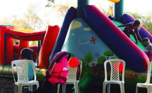 ילדים משחקים במגלשה מתנפחת צבעונית; כיסאות לבנים ועגלות תינוקות ניצבים בחזית התמונה.